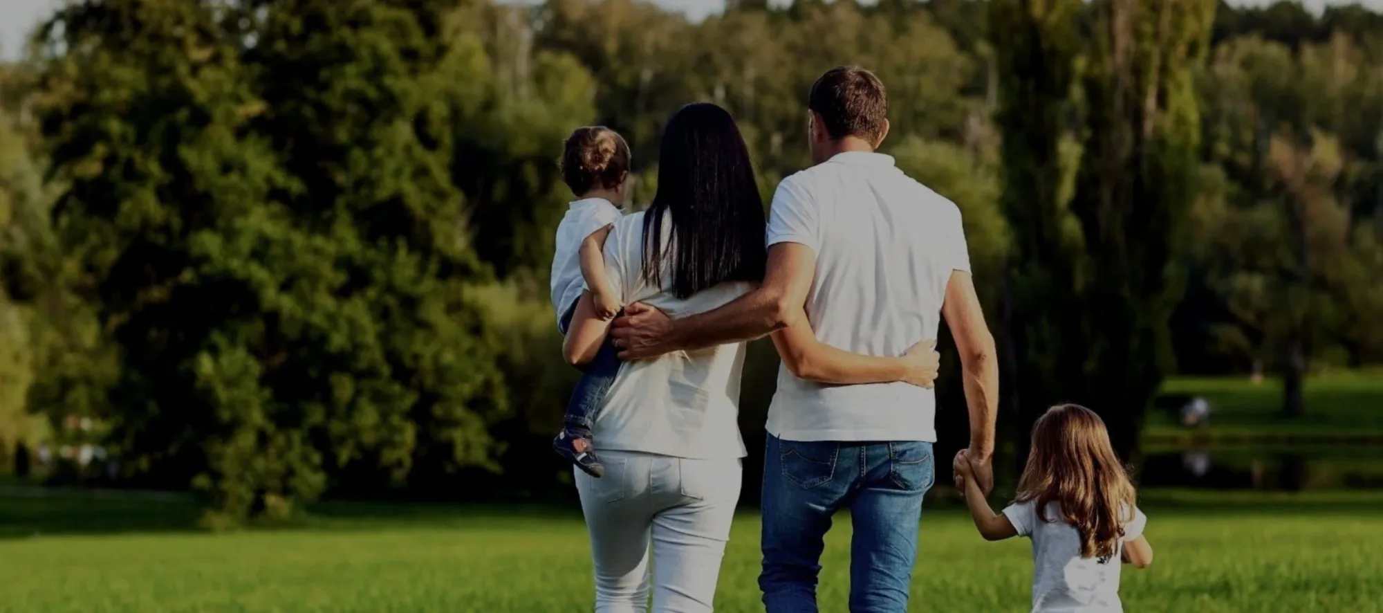 Family walking together across a sunny park lawn, seen from behind