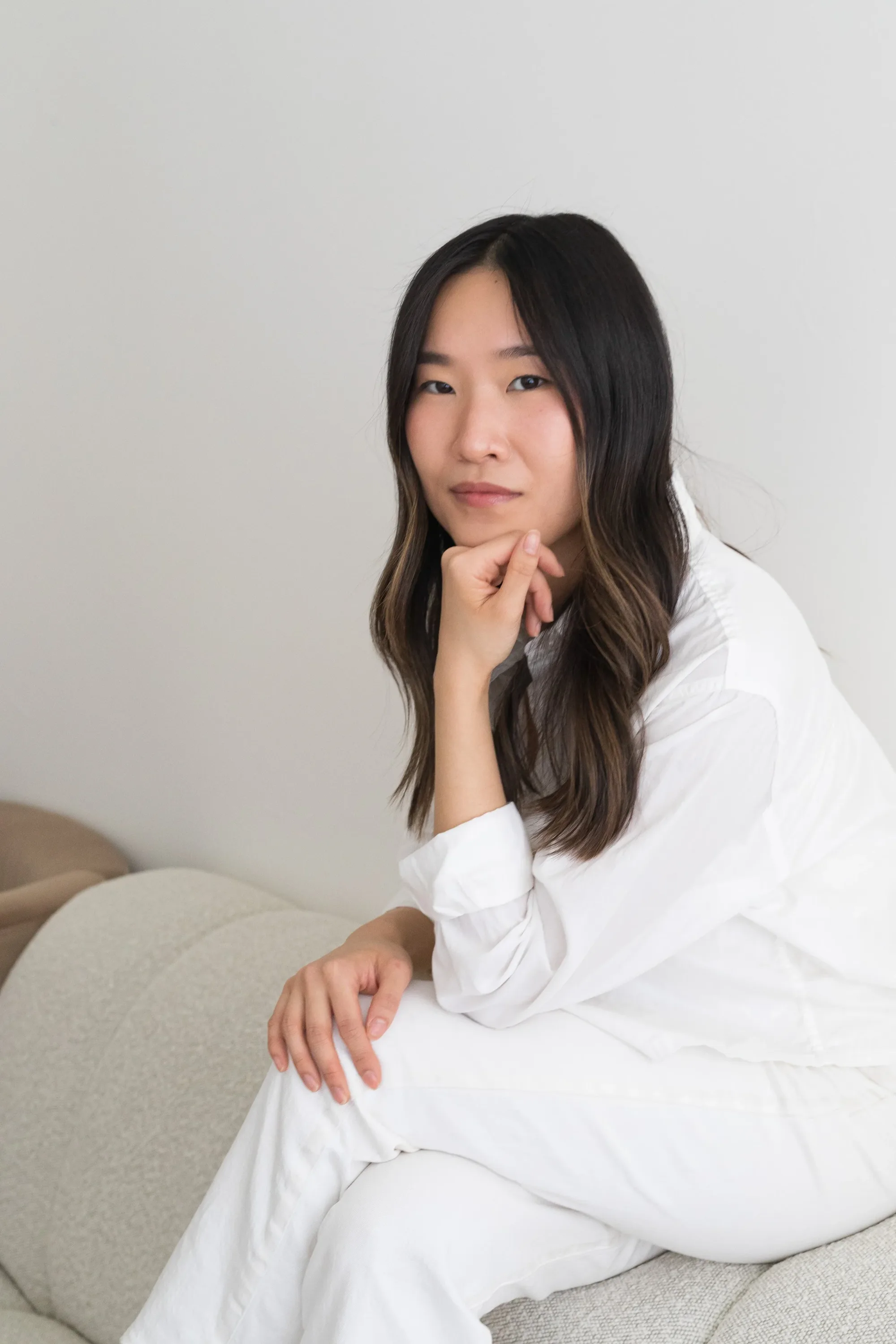 Woman in white seated with her chin resting on her hand in a bright room
