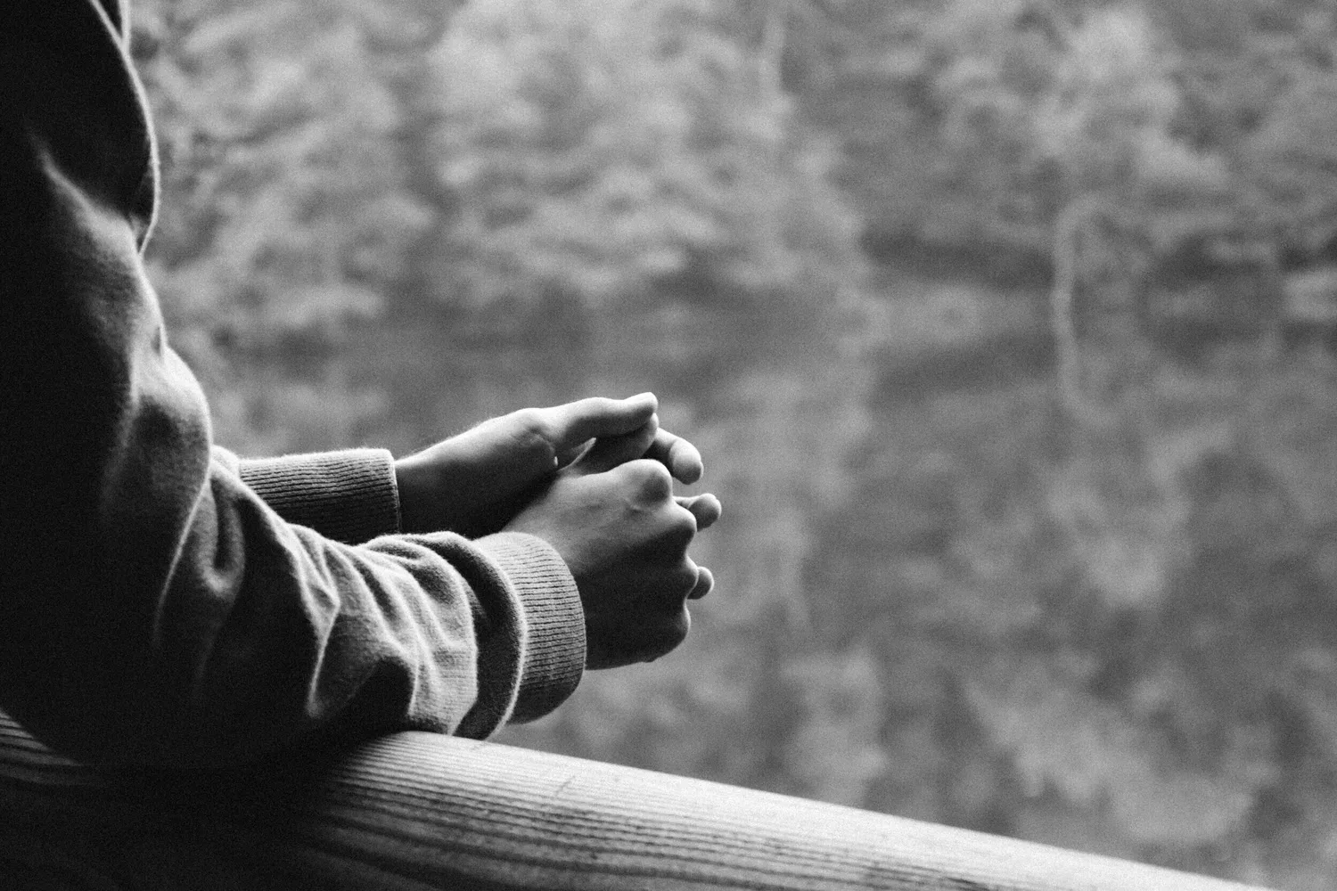 Black-and-white photo of hands clasped on a railing overlooking trees
