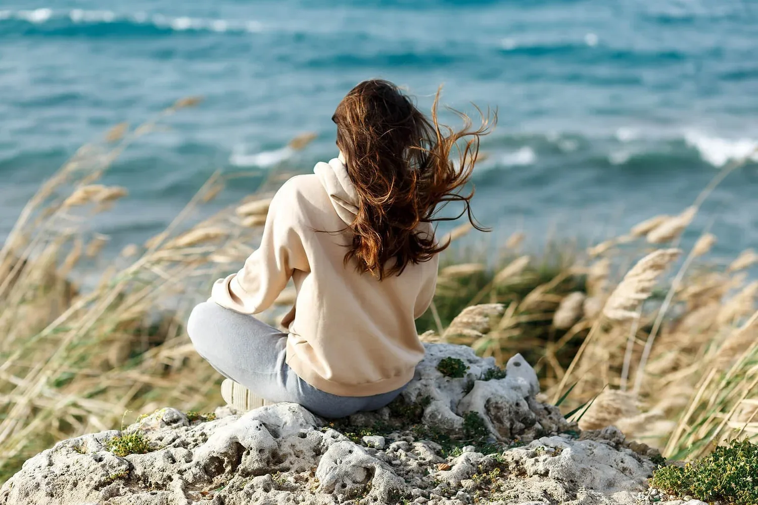 Woman sitting in tall grass by the sea, hair blowing in the wind
