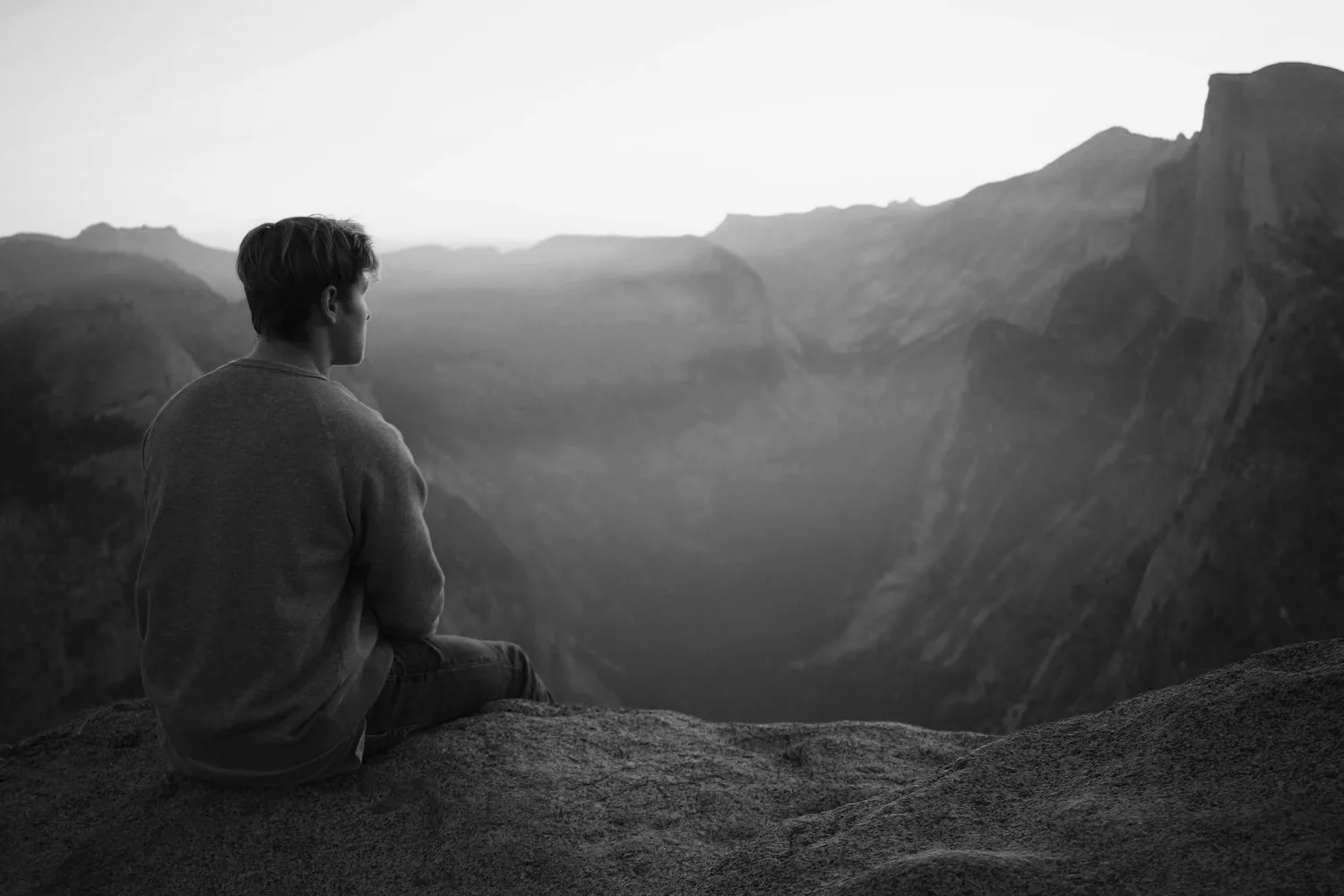 Black-and-white photo of a person sitting on a cliff edge overlooking hills