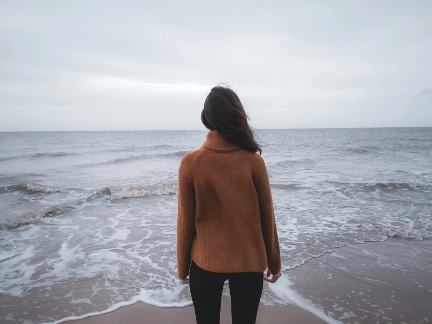 Woman in a brown coat standing on a beach facing the ocean