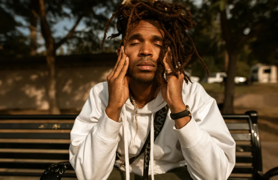 Young man in a white hoodie sitting on a bench outdoors, rubbing his temples