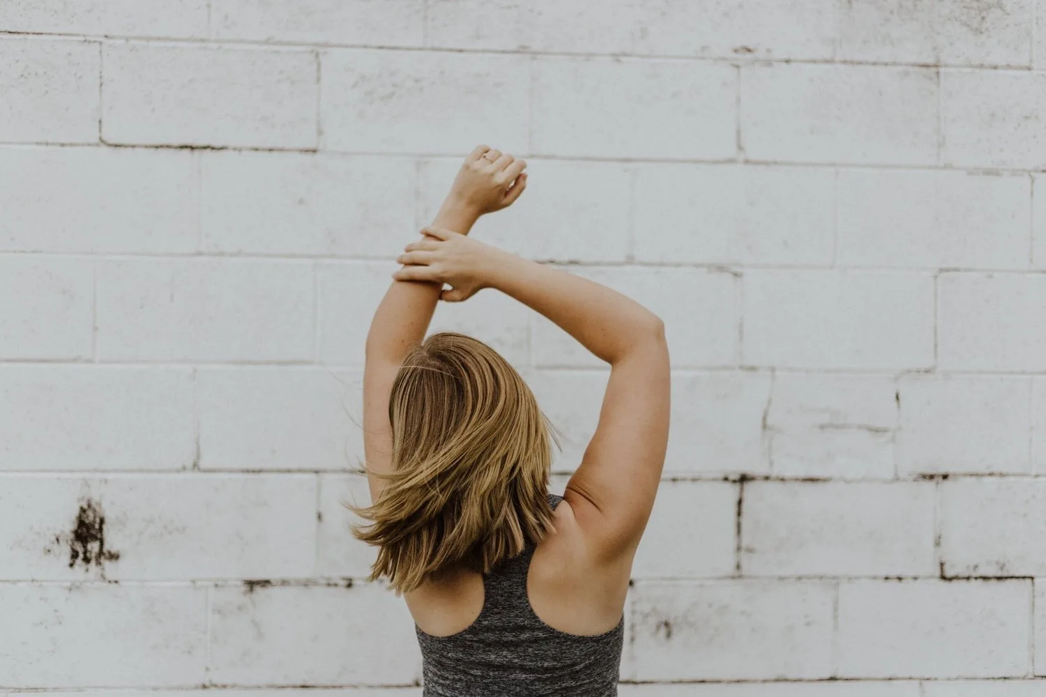 Person with arms raised overhead standing against a white brick wall