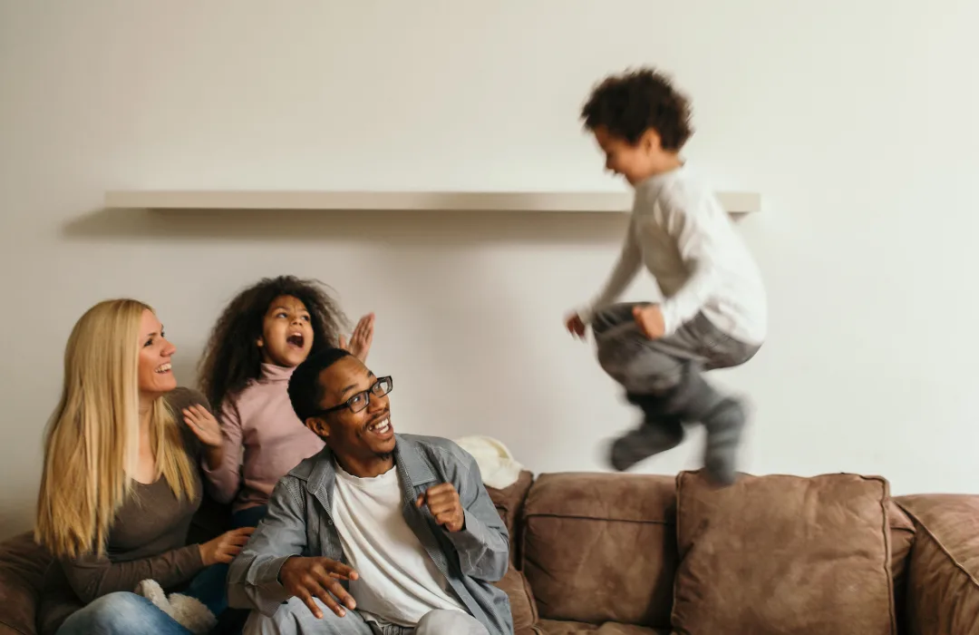 Parents talking on a sofa while their child jumps on the cushions