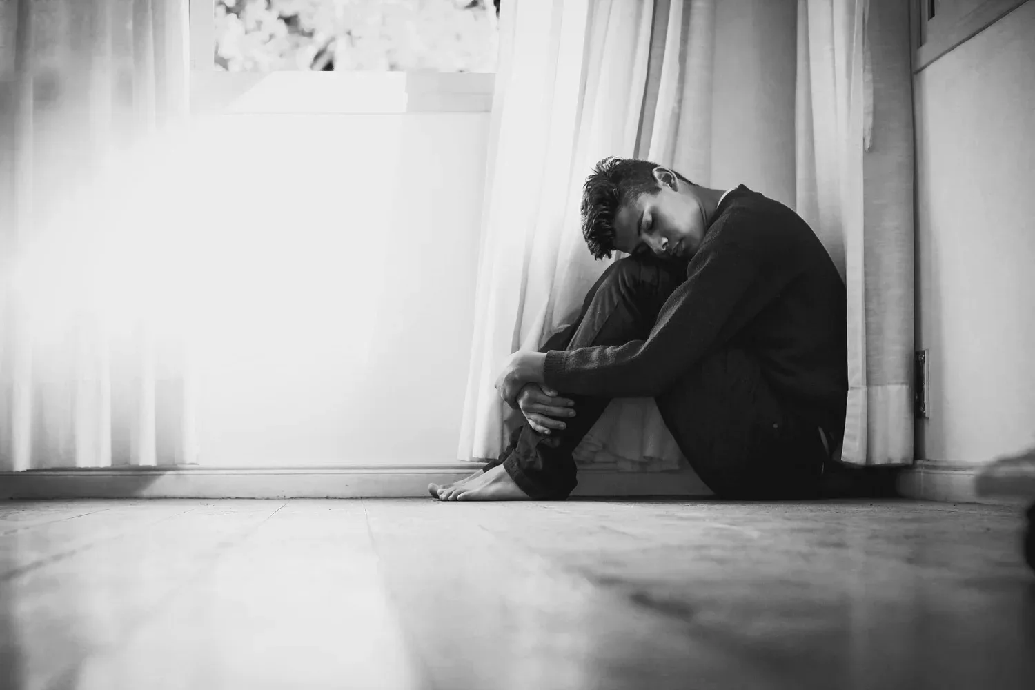 Black-and-white photo of a person sitting on the floor hugging their knees by a window