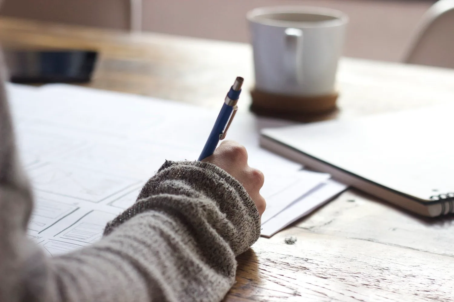 Hand writing with a pen in a notebook beside a mug