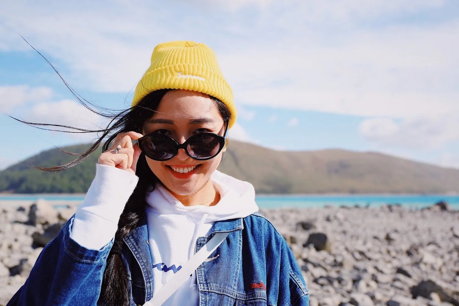 Young woman in a yellow beanie and sunglasses smiling on a rocky beach
