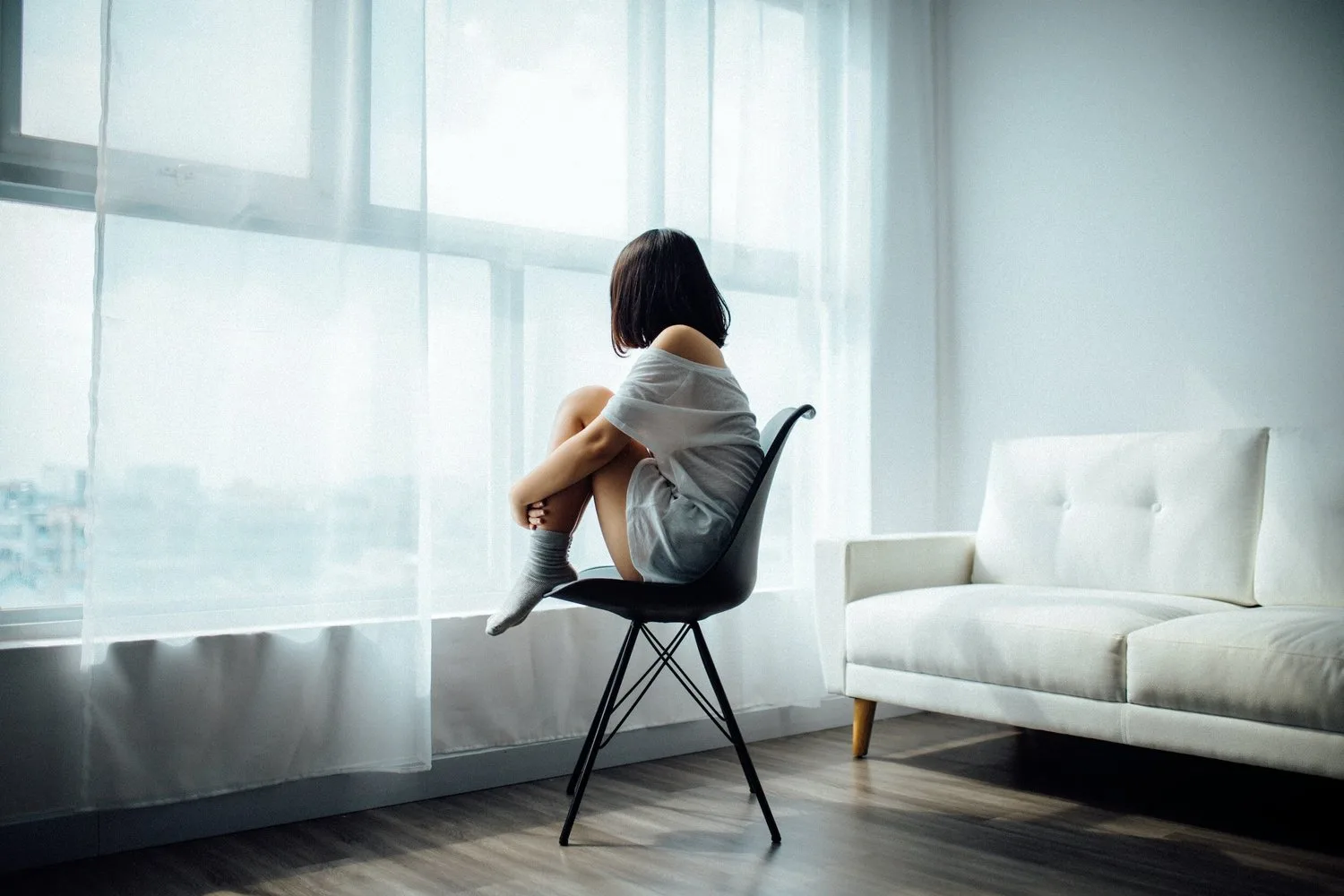 Person sitting alone on a chair facing a large window in a quiet room