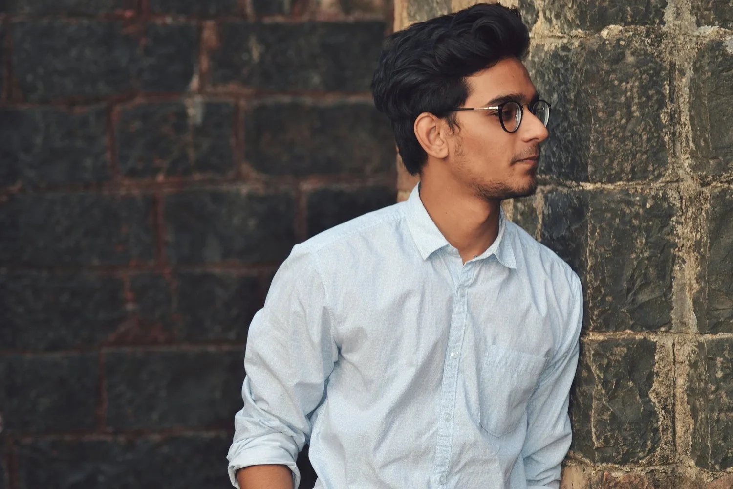 Young South Asian man in a white shirt looking to the side, standing by a dark wall