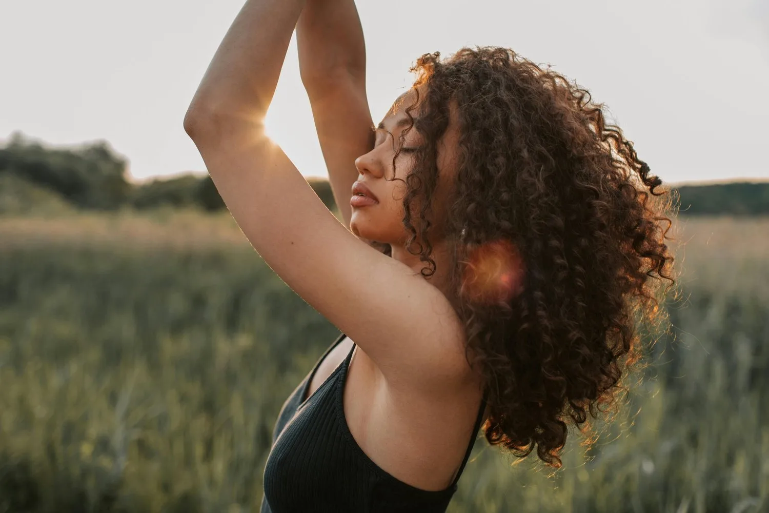 Woman with curly hair dancing with arms raised in a sunlit field