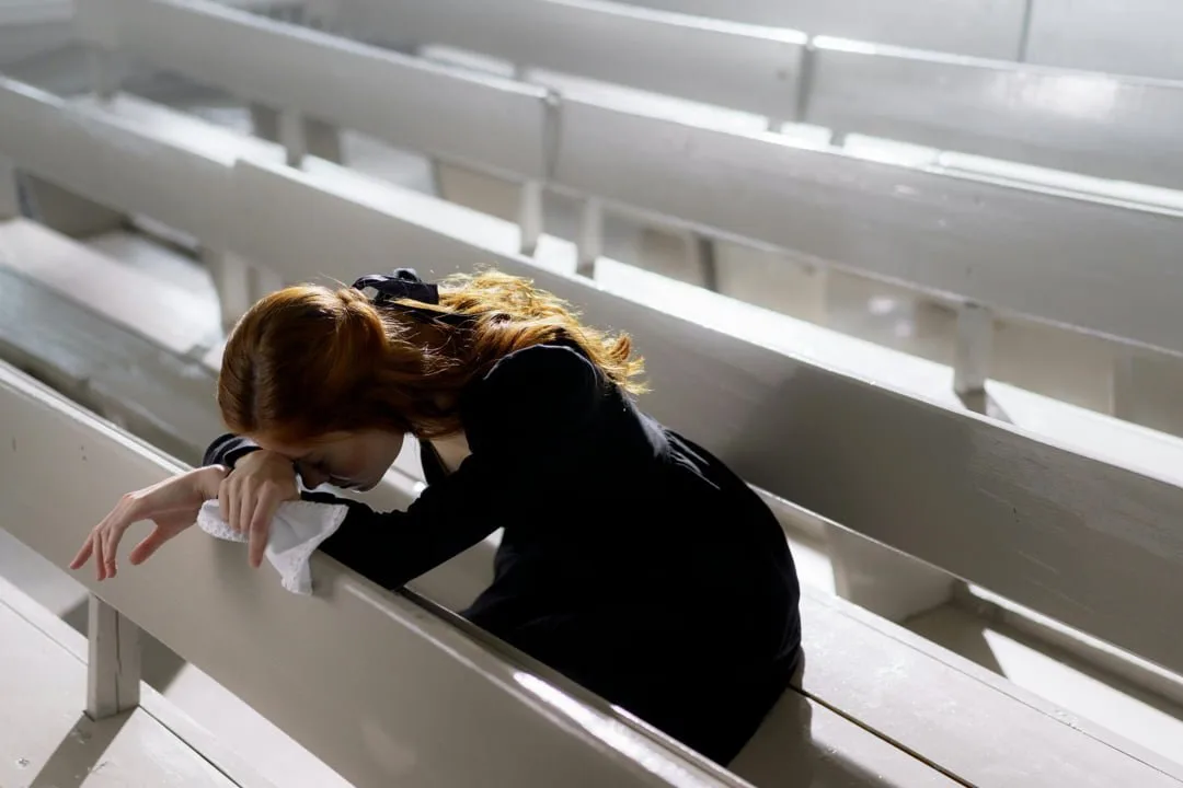 Person bent over with their head down between rows of white church pews