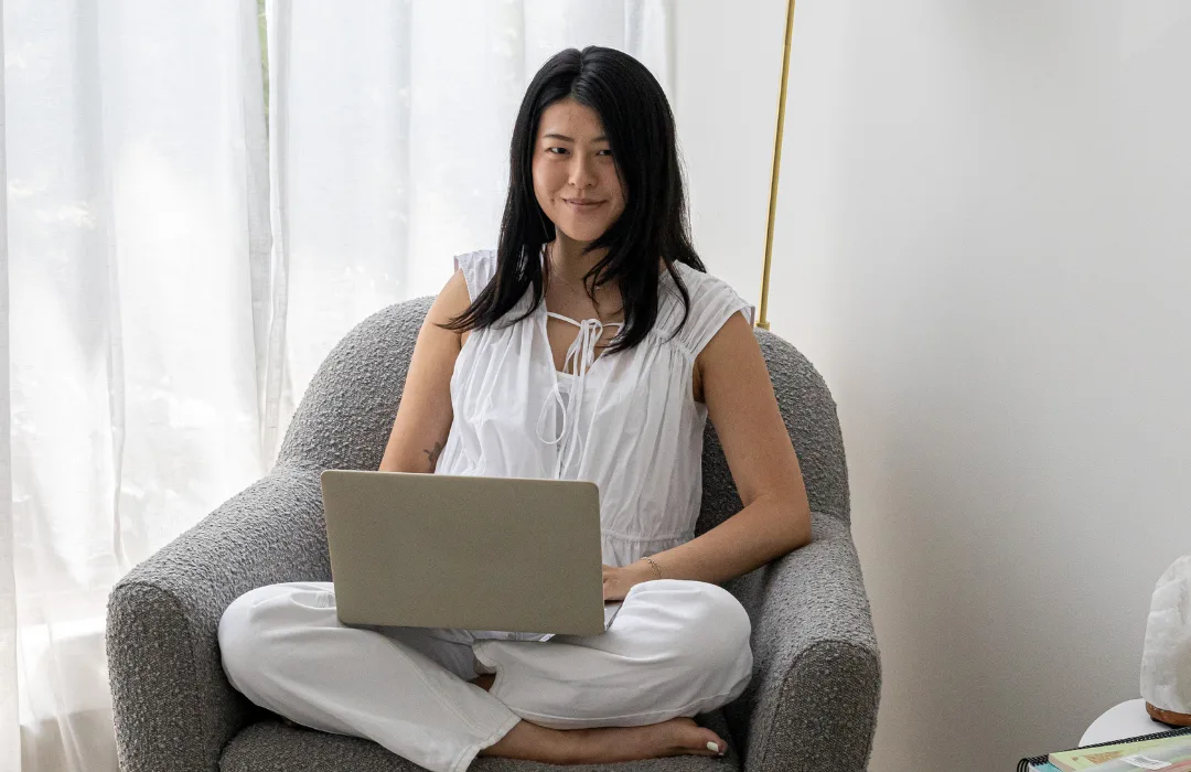 Woman in white sitting cross-legged in an armchair with a laptop