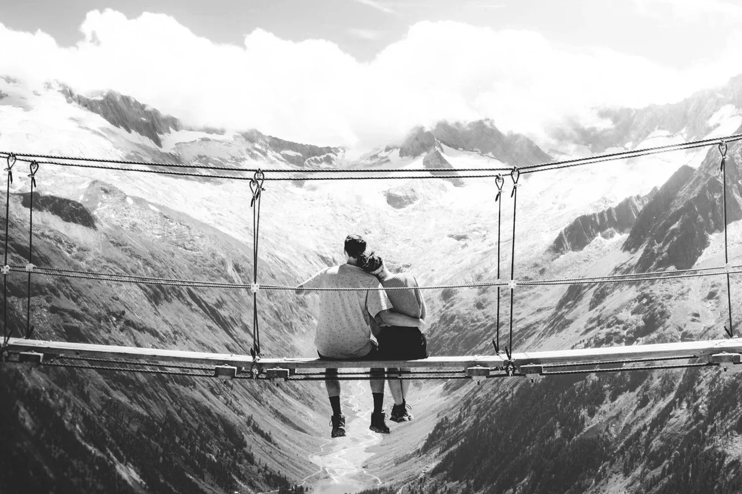 Black-and-white photo of two people sitting on a rope bridge in the mountains