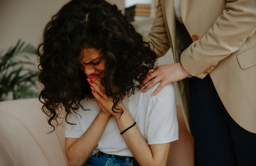 Tearful woman with curly hair covering her mouth as someone places a hand on her shoulder