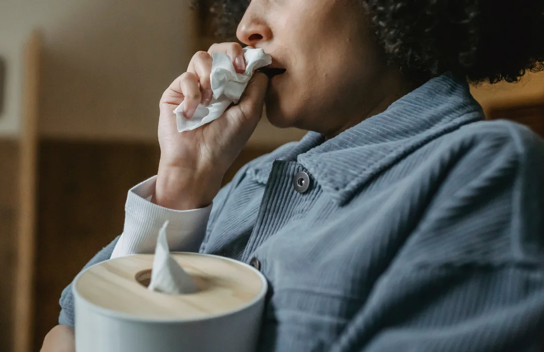 Person in a denim shirt covering their mouth with a tissue, seated beside a tissue box