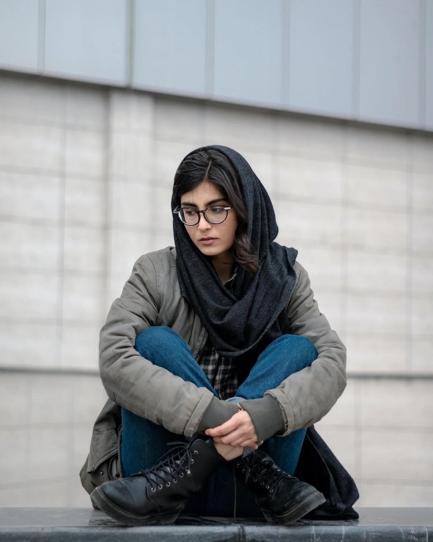 Woman in a hijab and glasses sitting outdoors hugging her knees