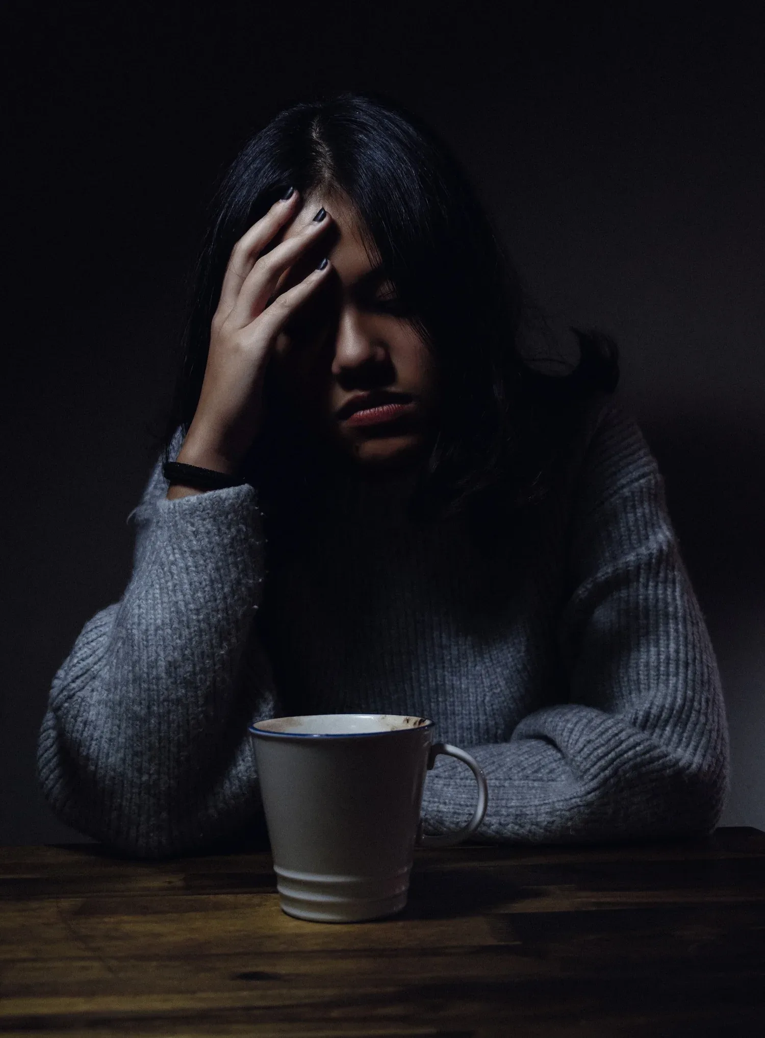 Person sitting in a dark room with a hand to their forehead beside a mug