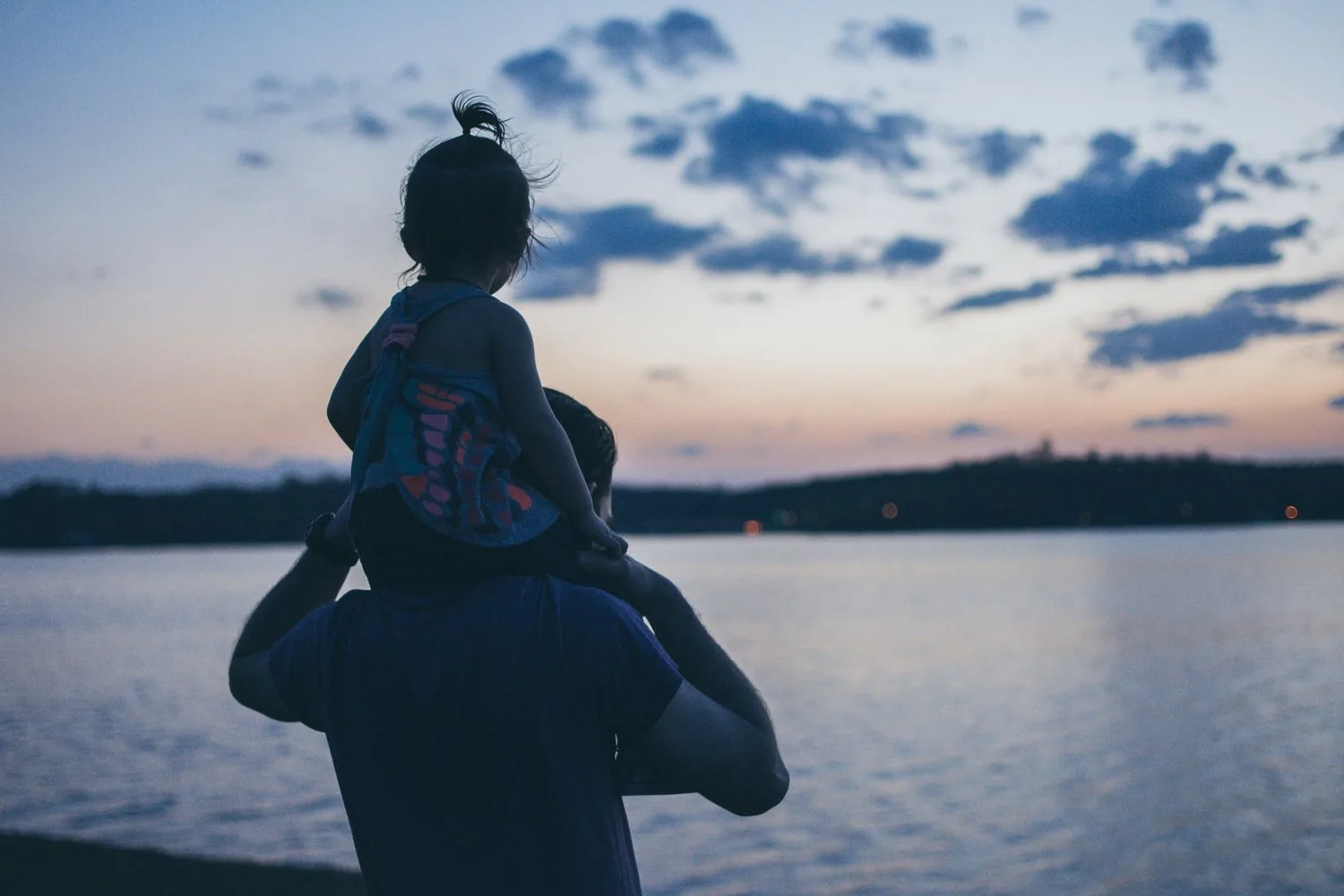 Silhouette of a child on a parent’s shoulders by a lake at dusk