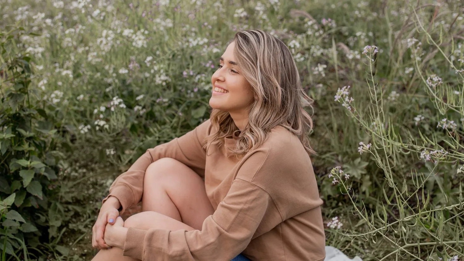 Woman sitting in tall grass with her arms around her knees, smiling