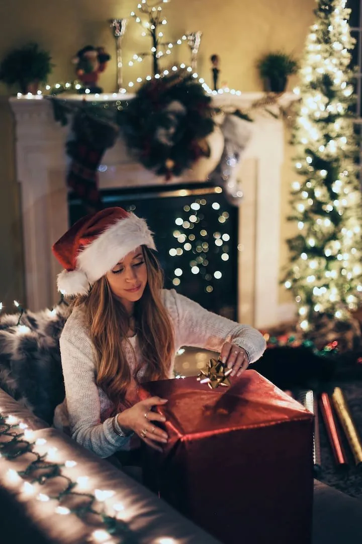 Young woman in a Santa hat opening a gift beside a lit fireplace and tree