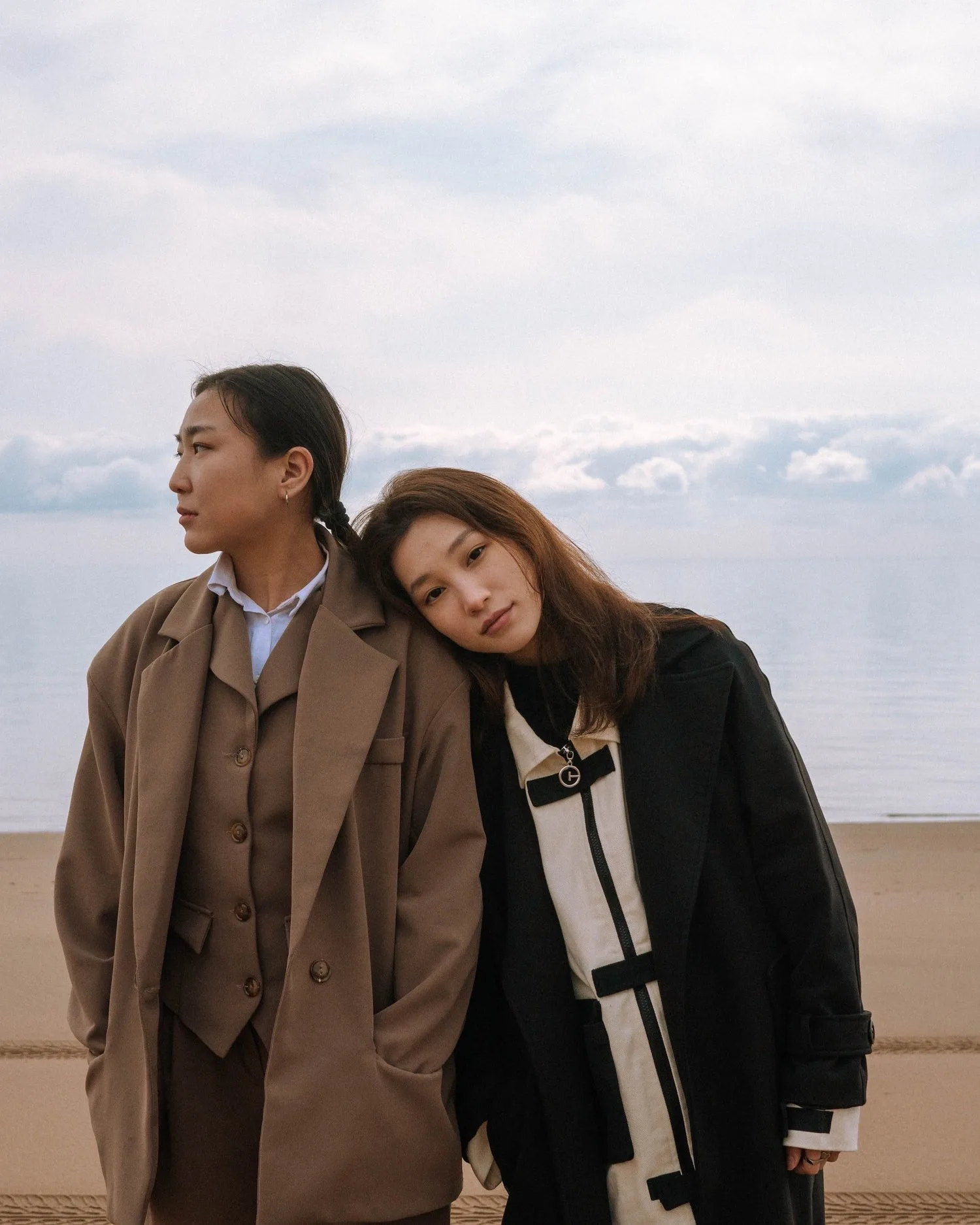 Two women standing close together by the sea, one resting her head on the other’s shoulder