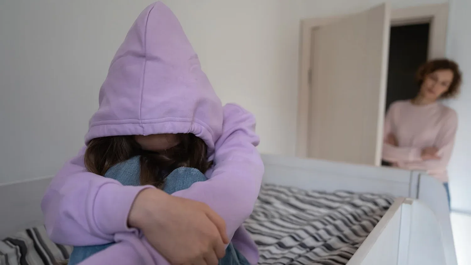 Person in a purple hoodie sitting hunched on a bed while someone looks in from the doorway