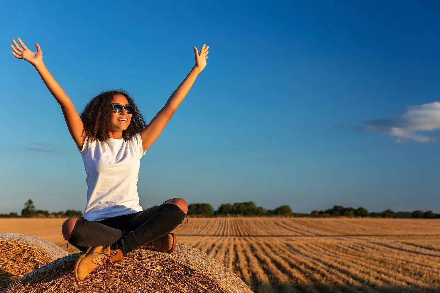 Woman sitting in a field with arms raised joyfully under a blue sky