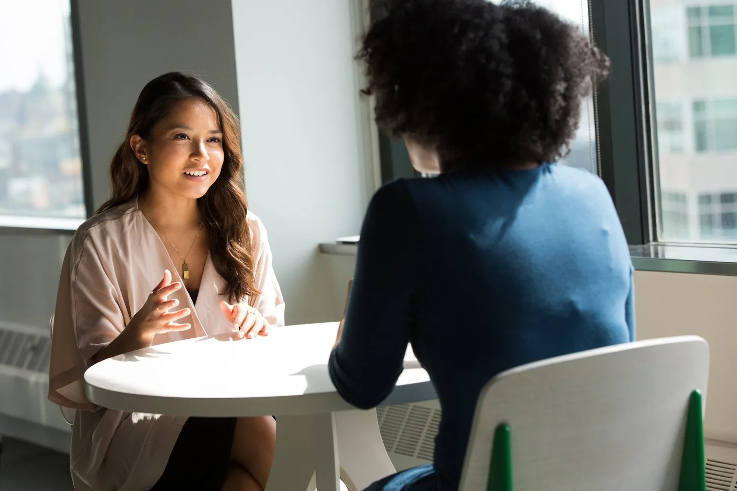Two women talking across a small round table in a sunny room