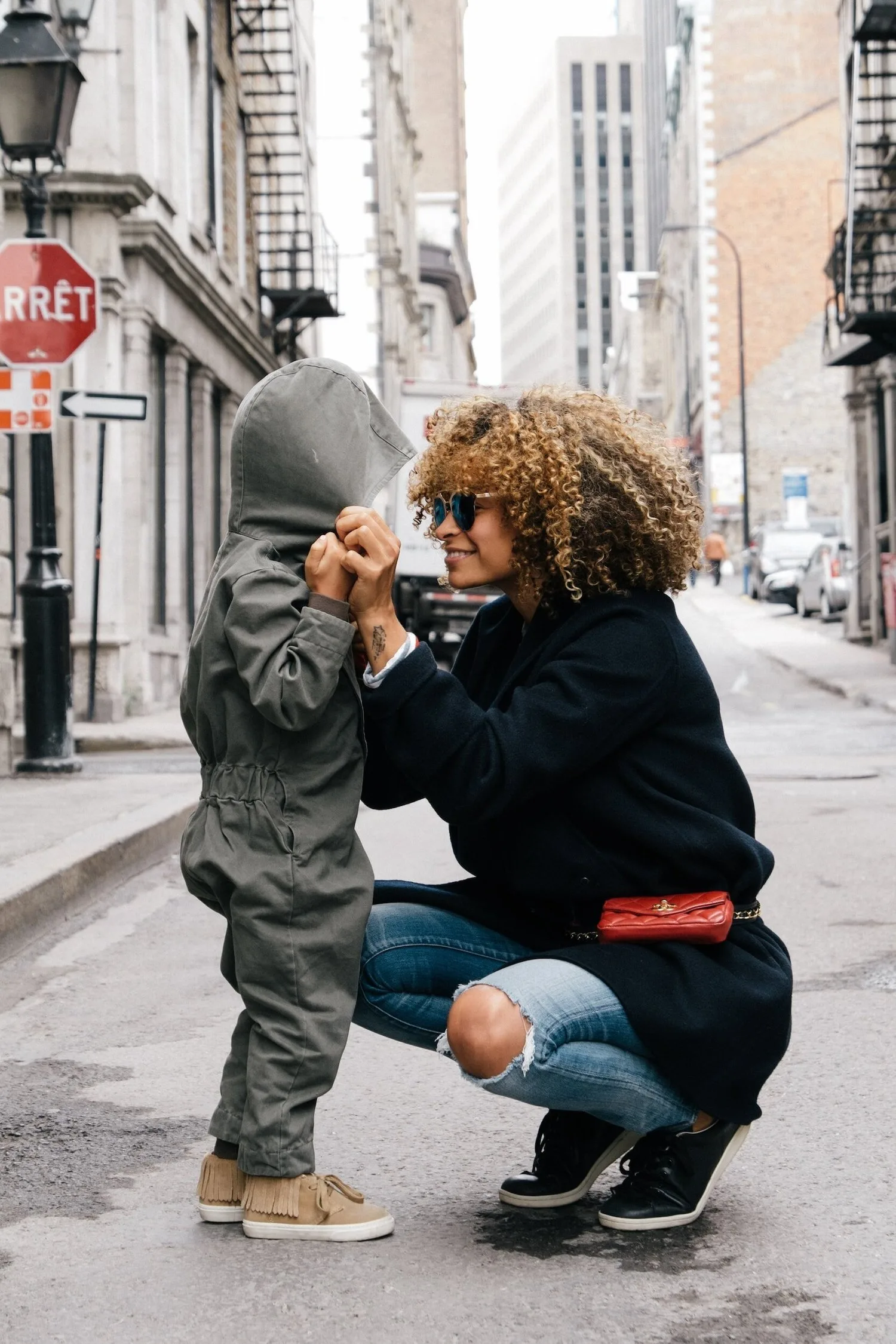 Mother kneeling on a city sidewalk to adjust her toddler’s jacket