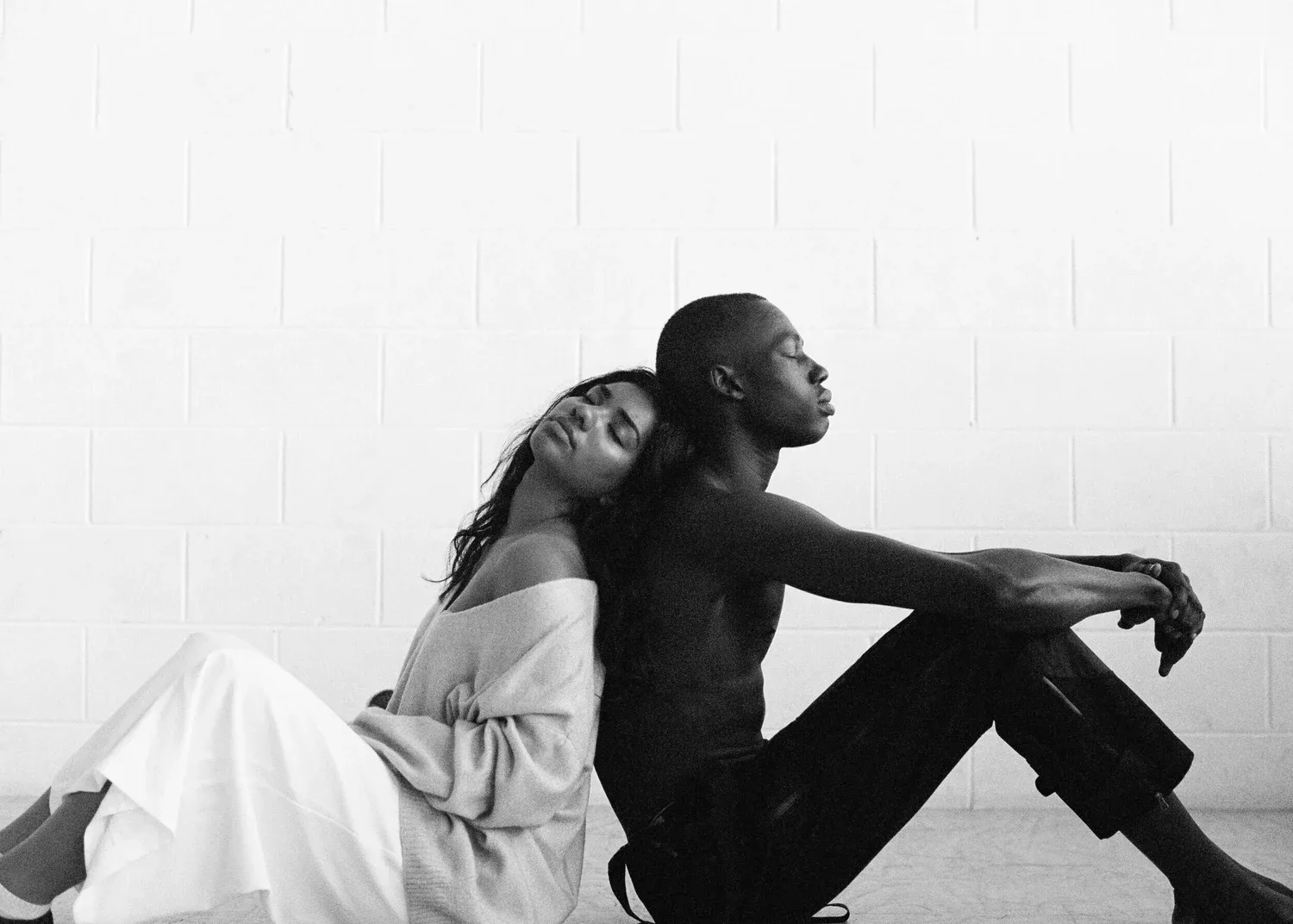 Black-and-white photo of two people sitting back to back on the floor