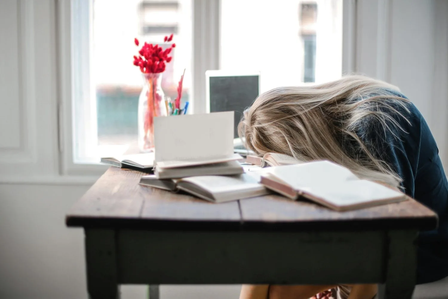Person slumped over a desk with an open book beside a window