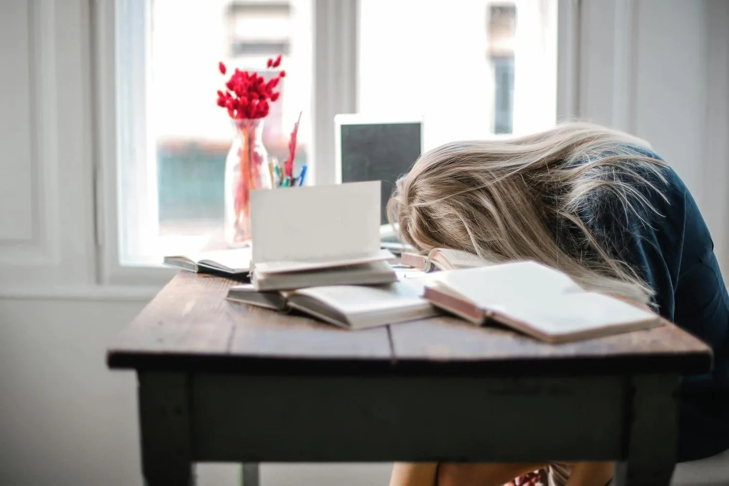 Person slumped over a desk with an open book beside a window