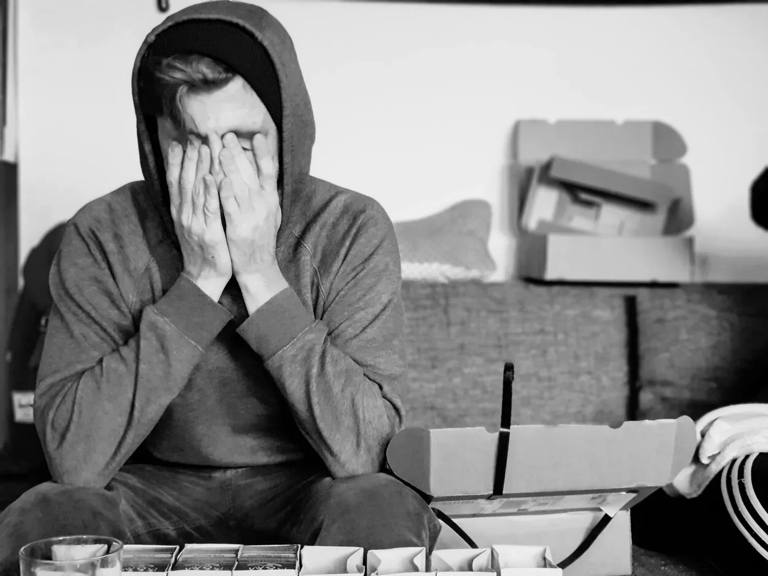 Black-and-white photo of a person in a hoodie covering their face beside moving boxes