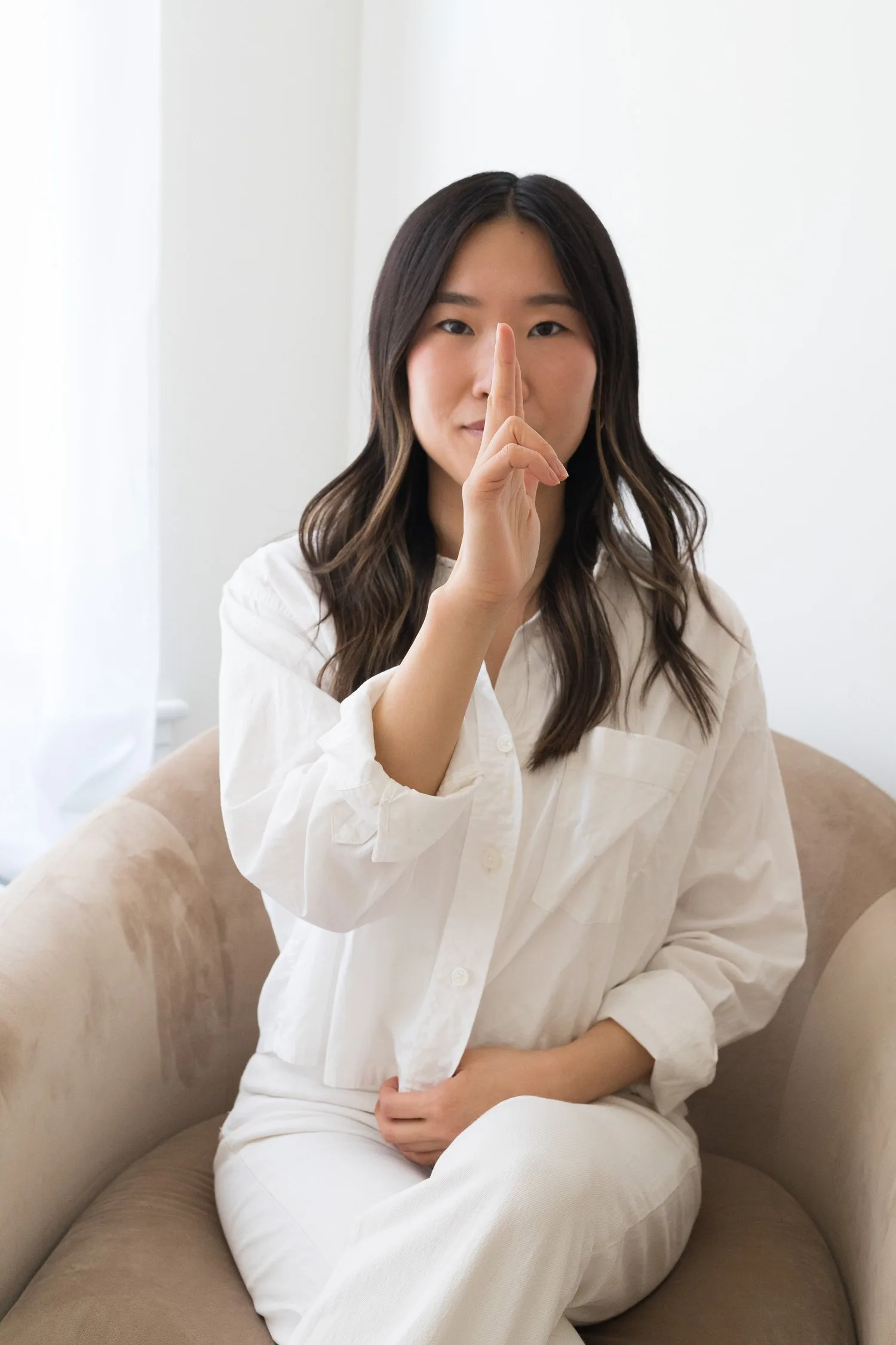 Woman in white seated in an armchair holding one finger in front of her face