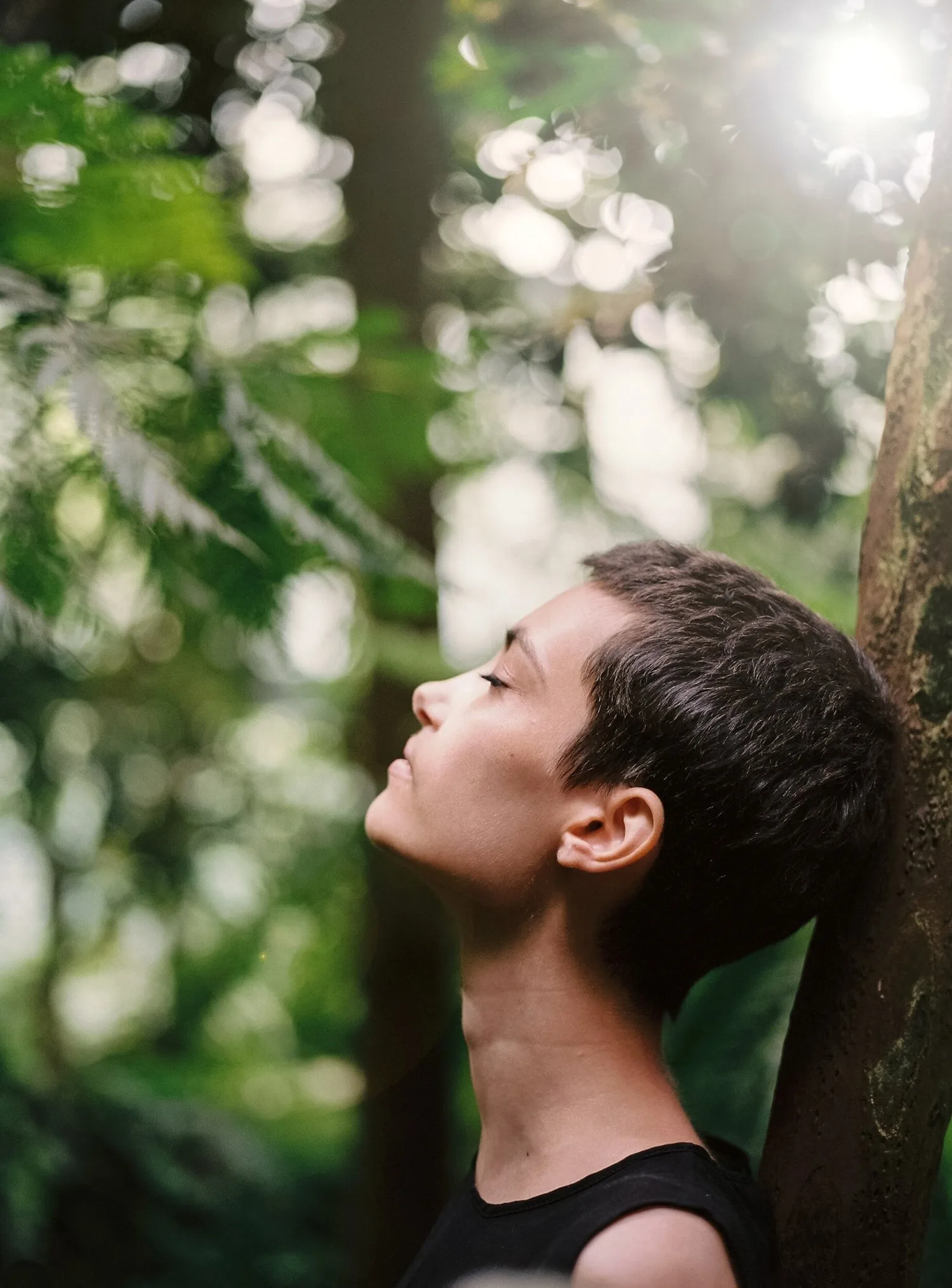 Person with short hair tilting their face toward sunlight among trees