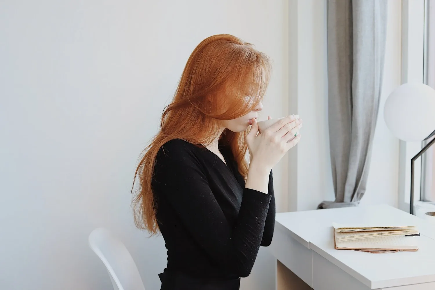 Woman with red hair sipping from a mug by a bright window