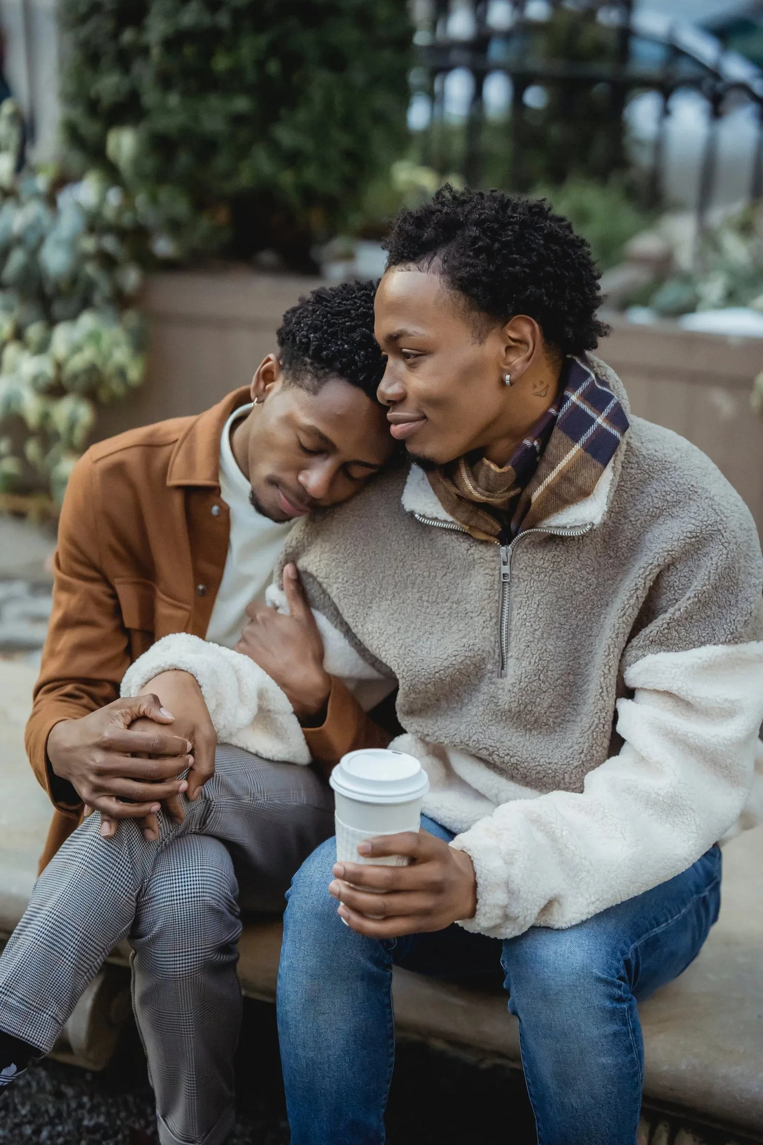 Two people sitting close together outdoors, one resting a head on the other’s shoulder