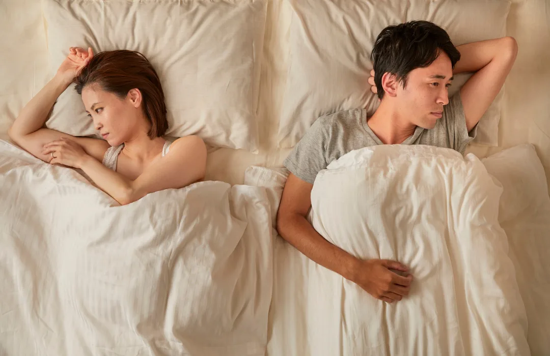 Two people lying awake in bed under white sheets, facing away from each other