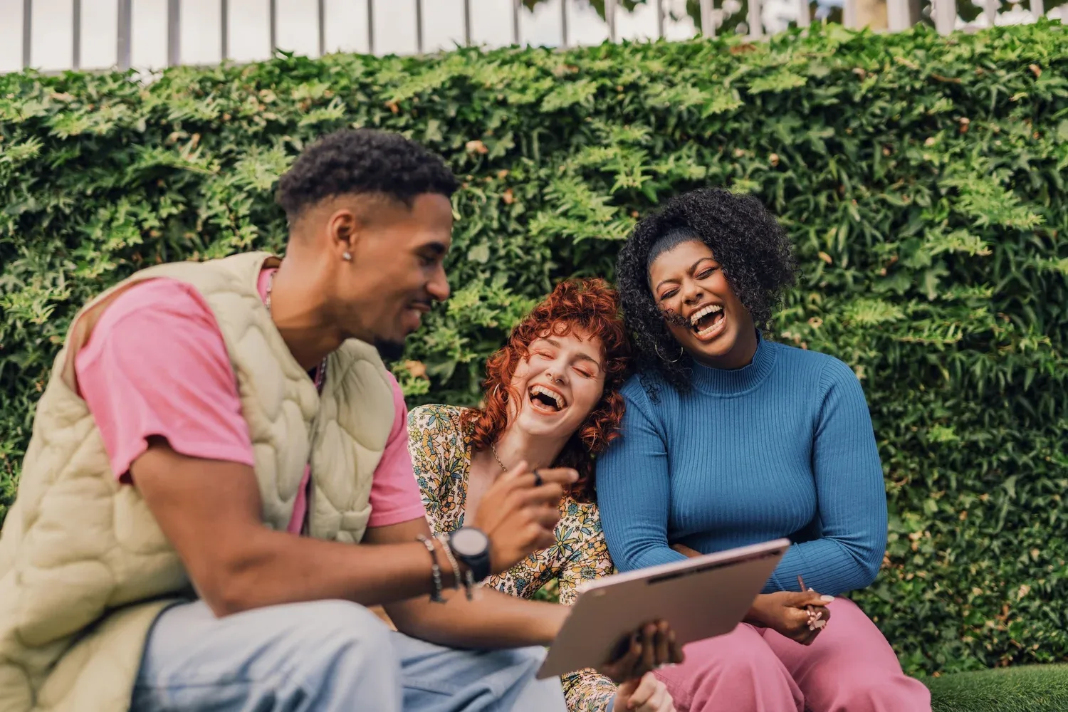 Three friends laughing together on a bench in front of a hedge