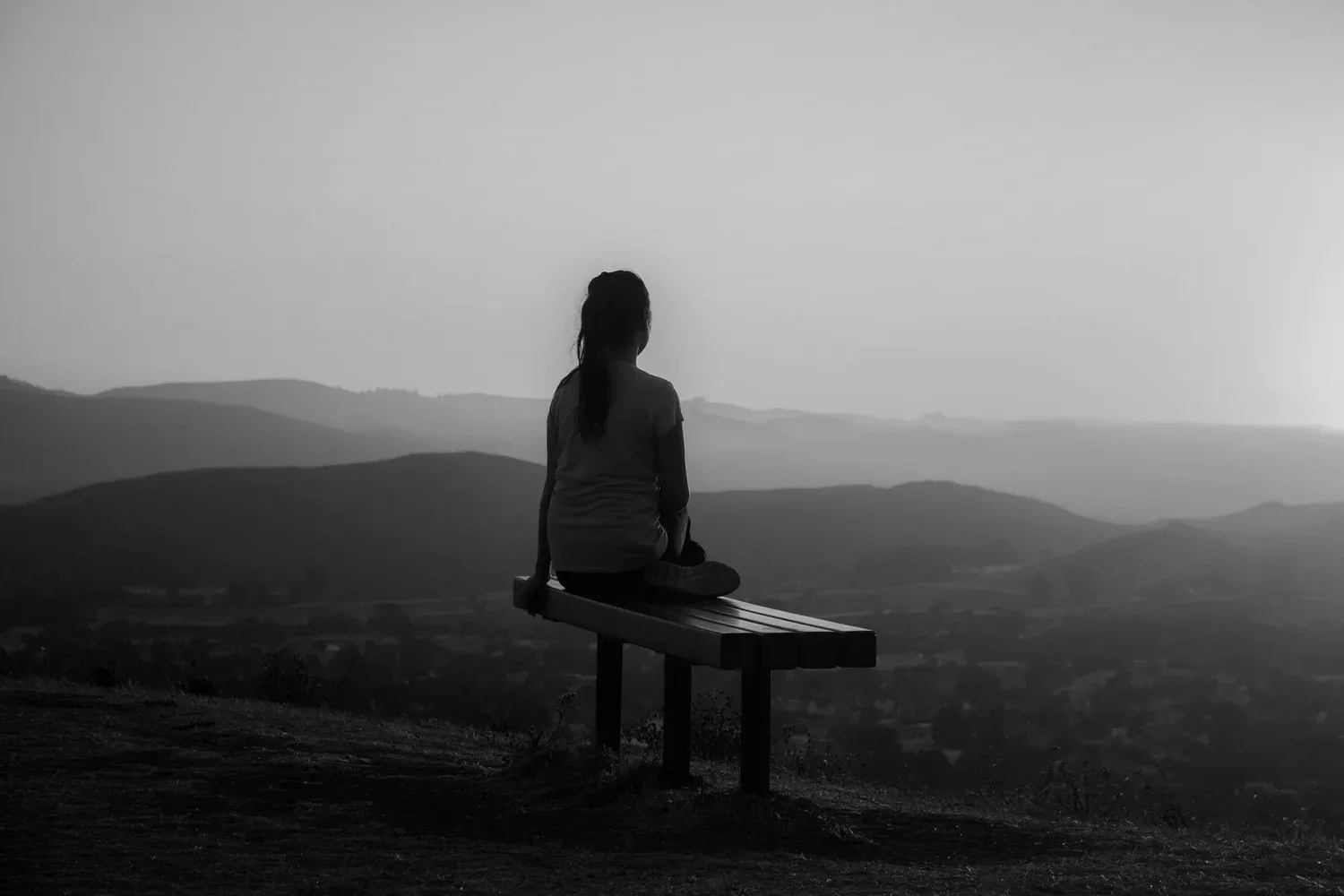 Black-and-white photo of a person sitting on a bench overlooking misty hills