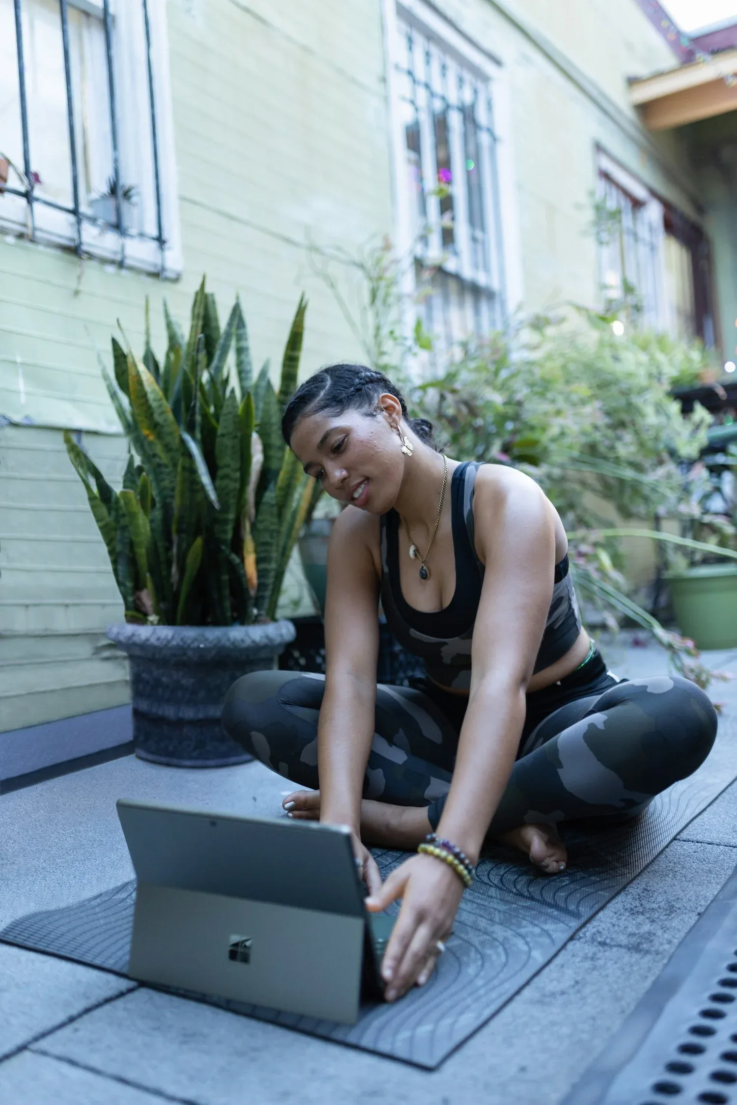 Woman sitting on a balcony floor typing on a laptop beside plants
