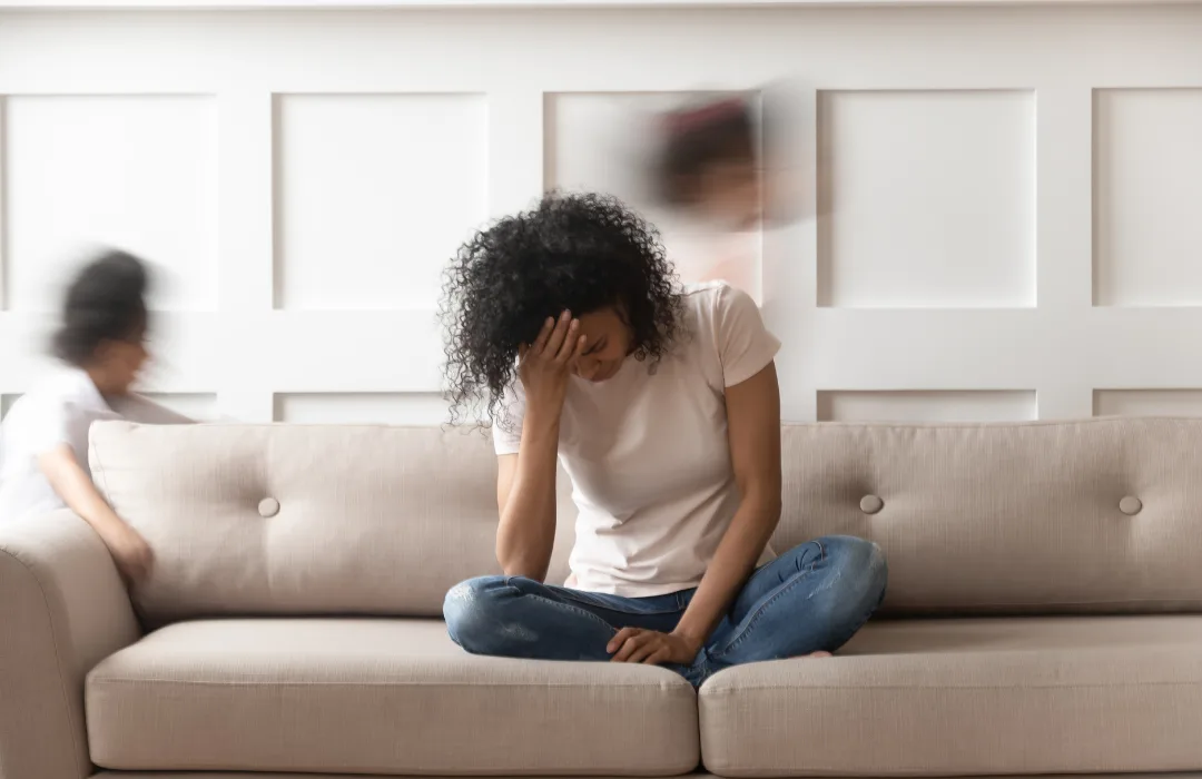 Woman sitting on a sofa with her head down while blurred figures move around her