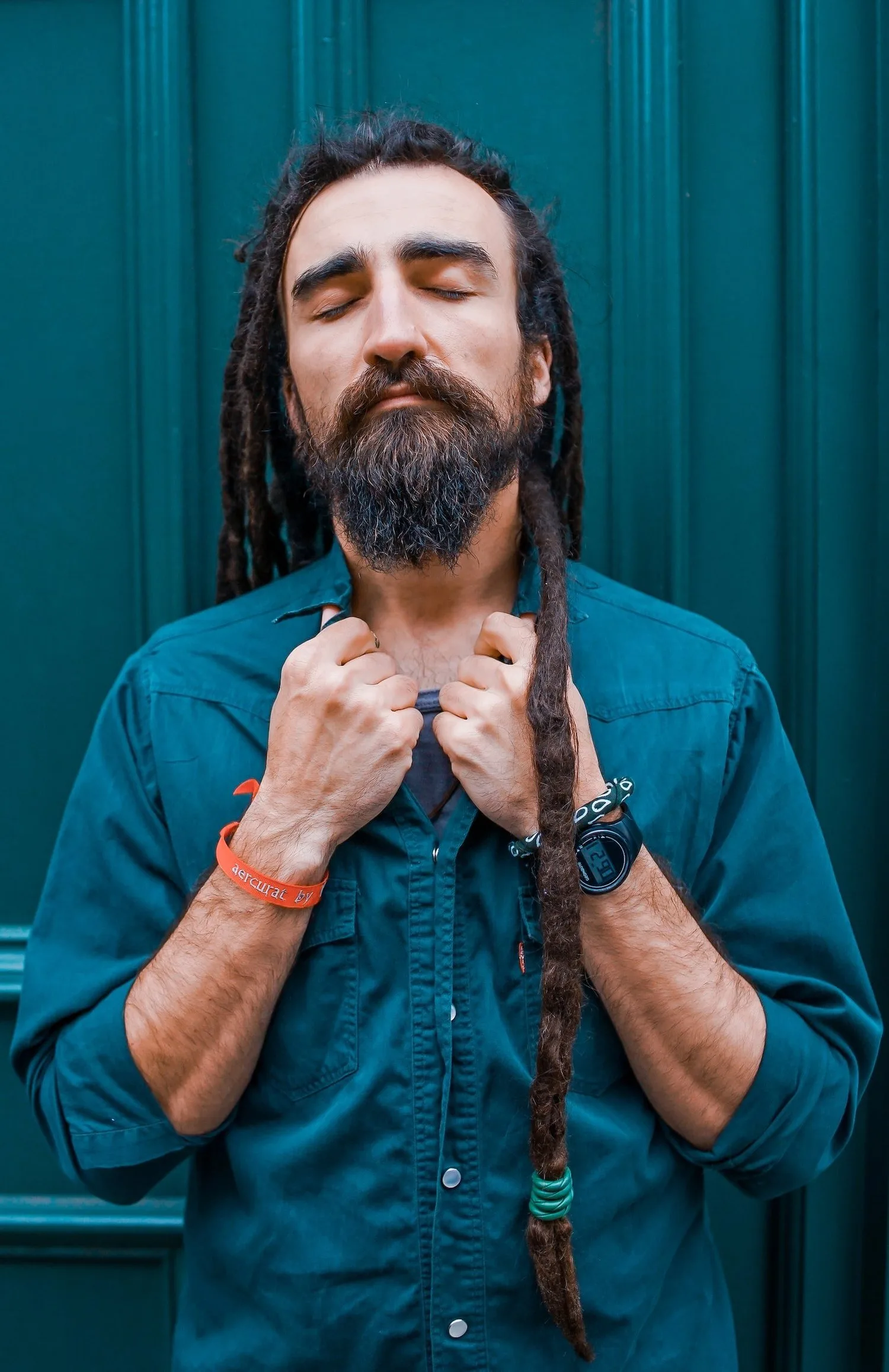 Man with long locs and a beard in a denim shirt, hands at his chest