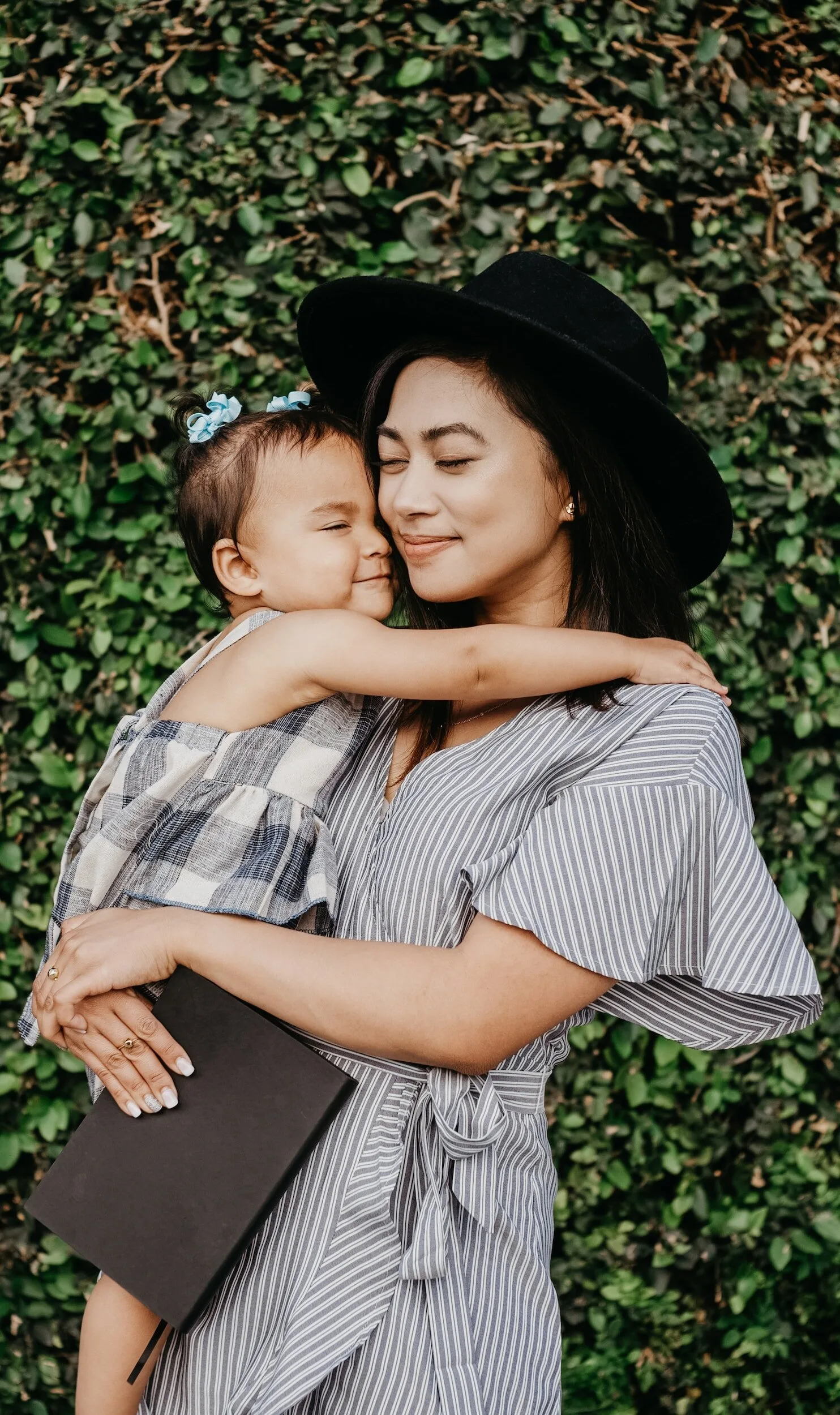 Smiling mother in a wide-brimmed hat holding her baby in front of greenery