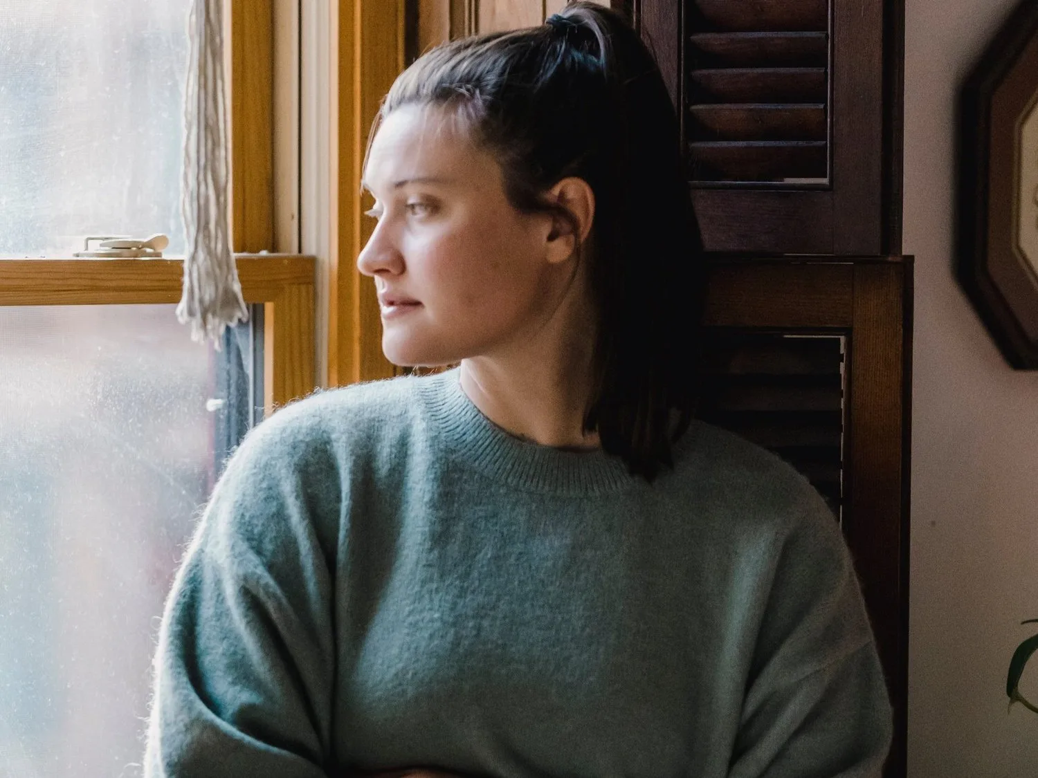 Woman in a gray sweater gazing out a window, looking thoughtful