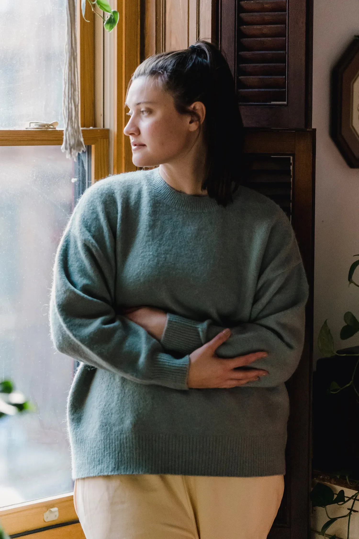 Woman in a gray sweater standing by a window with arms folded across her middle