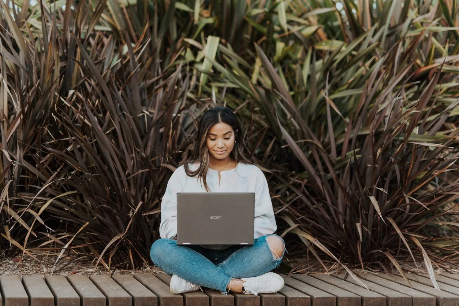 Woman sitting cross-legged outdoors with a laptop in front of tall grasses
