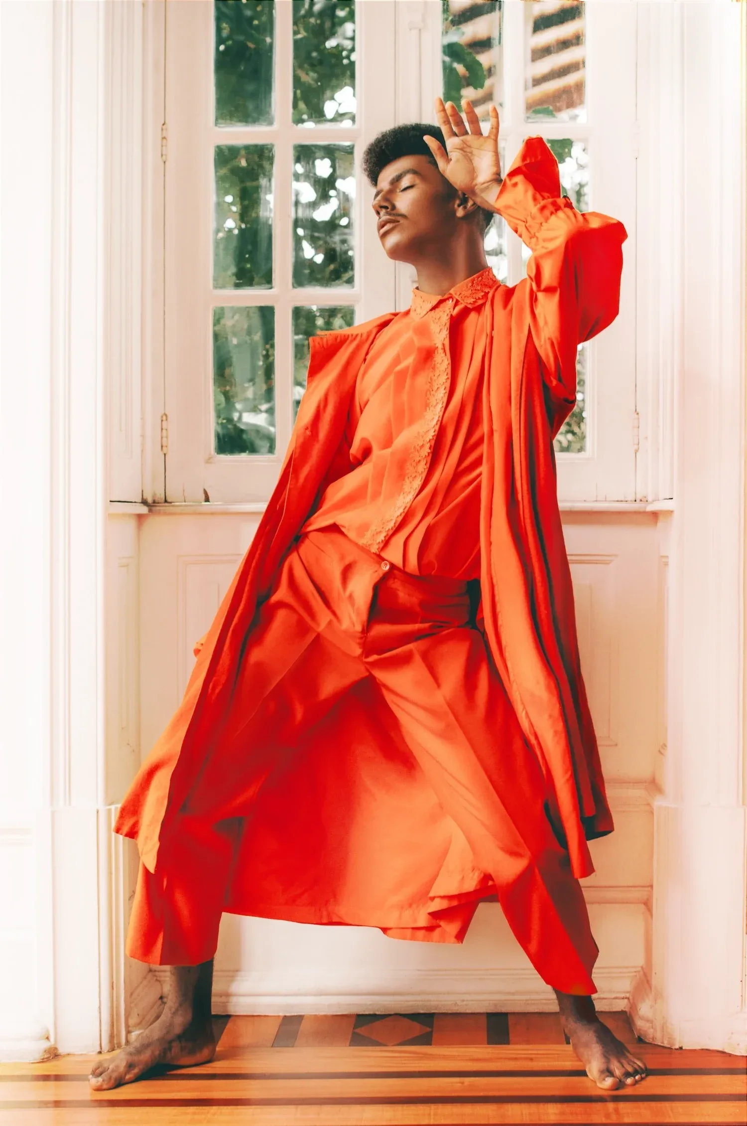Person in a flowing orange outfit posing with arms raised in a sunlit doorway