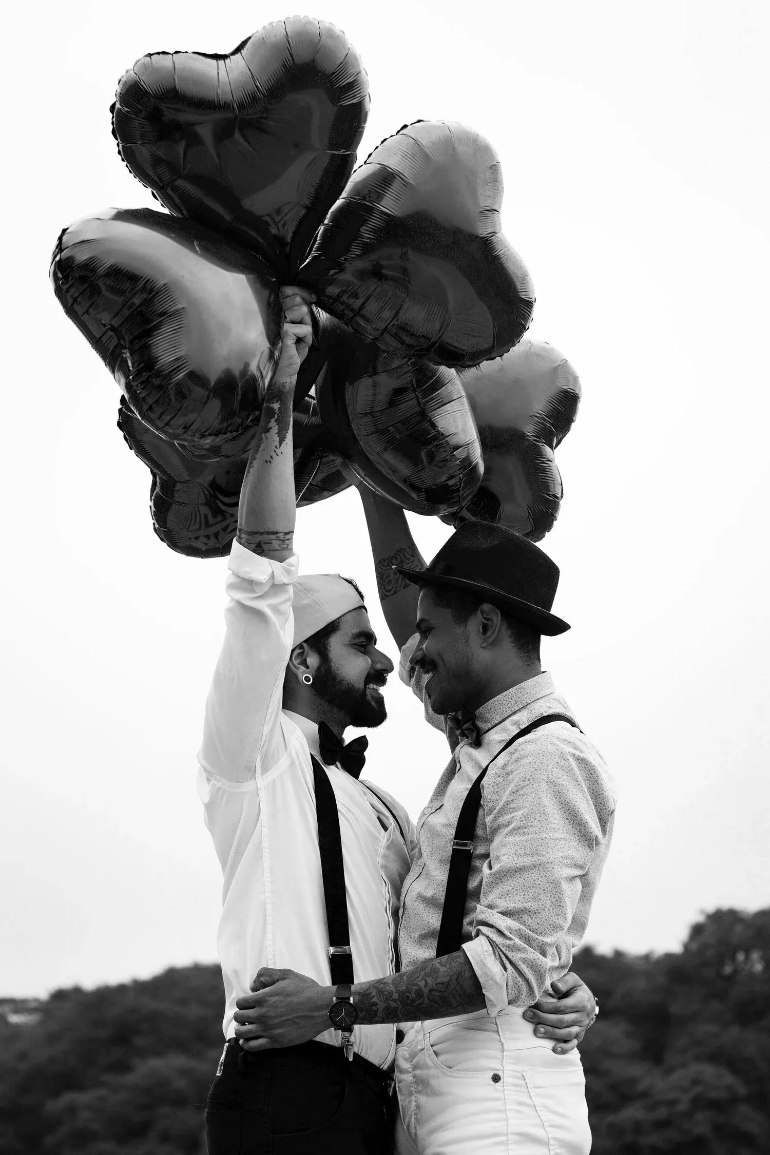 Black-and-white photo of two men embracing while holding heart-shaped balloons